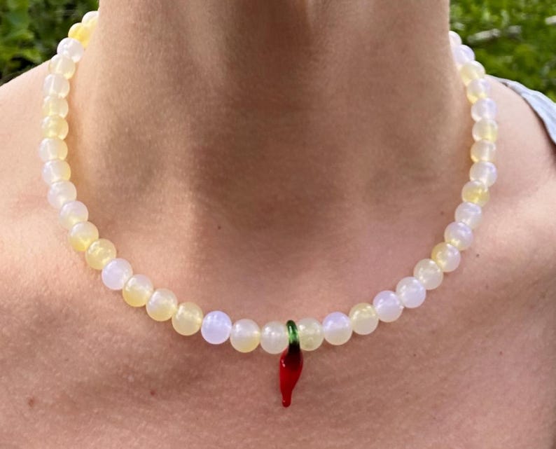 May include: A beaded necklace with a pendant. The necklace features a strand of round, light yellow and white beads. A small, red chili pepper pendant with green accents hangs from the center of the necklace.