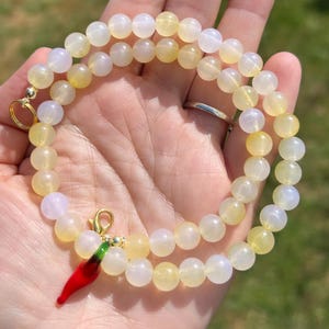 May include: A beaded necklace featuring round, pale yellow and white beads. The necklace has a gold-colored clasp and a red chili pepper charm with green accents. The necklace is displayed on a hand.