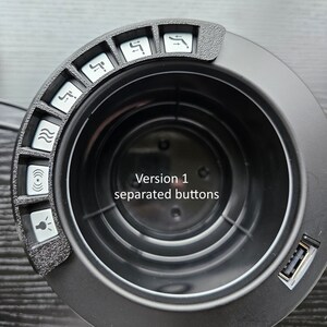 May include: Black circular device with multiple buttons and a USB port. The buttons have different symbols, including a wave, a light bulb, and a speaker. The text "Version 1 separated buttons" is visible on the device.