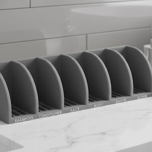 May include: A gray organizer with six labeled compartments for bathroom essentials. The labels read: Shampoo, Conditioner, Face, Body, Shave, and Scrub. The organizer is on a white countertop.