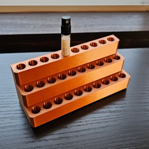 May include: A three-tiered orange plastic organizer with 24 round holes for storing small bottles. A small clear glass bottle with a black spray nozzle is in one of the holes. The bottle has a white label with the word "ALAR" printed on it.