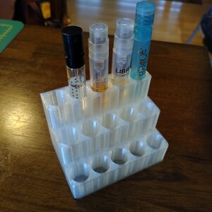 K&ouml;nnte beinhalten: Ein wei&szlig;er Plastik-Organizer mit mehreren F&auml;chern zum Aufbewahren von kleinen Flaschen. Vier kleine Flaschen sind im Organizer ausgestellt, zwei sind klar mit brauner Fl&uuml;ssigkeit, eine ist klar mit blauer Fl&uuml;ssigkeit und eine ist schwarz mit einem silbernen Etikett, auf dem "Narciso Rodriguez For Her" steht.