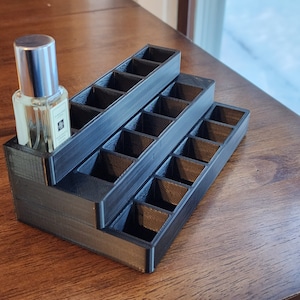May include: A black plastic tiered organiser with 18 compartments for storing small items, such as perfume bottles. A clear glass bottle with a silver cap is in one of the compartments.