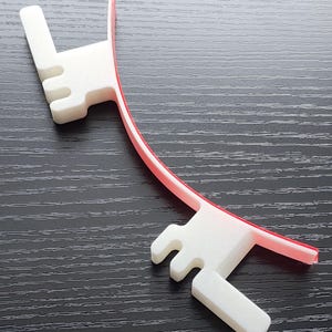 May include: A white plastic 3D printed object with a red plastic strip attached to it. The object has a curved shape and two prongs on each end.