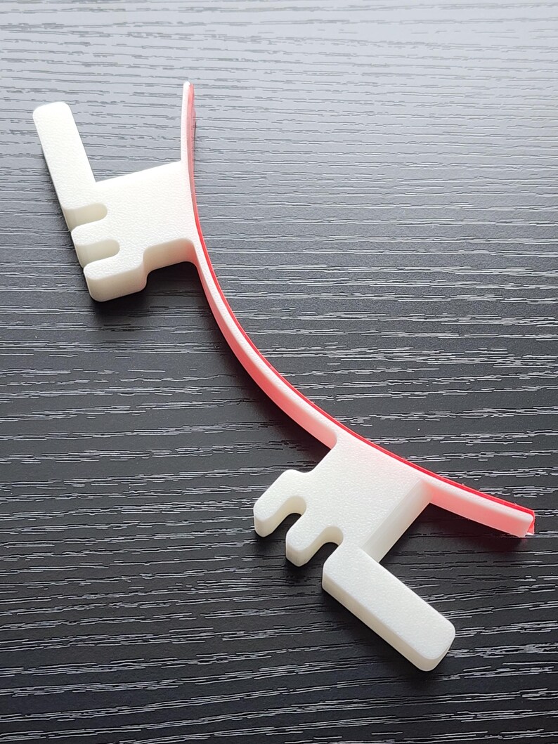 May include: A white plastic 3D printed object with a red plastic strip attached to it. The object has a curved shape and two prongs on each end.