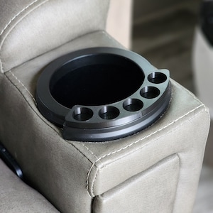 May include: A black, circular cup holder with five smaller, circular compartments. The cup holder is set into the armrest of a beige upholstered chair. The design is modern and functional, perfect for holding drinks and small items.