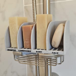 Può includere: Un porta sapone da doccia grigio con quattro scomparti per saponette. Gli scomparti sono etichettati "Shampoo", "Balsamo", "Corpo" e "Viso".