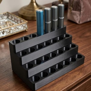 May include: A black tiered display stand with multiple circular cutouts for organizing small cylindrical items. Four metallic tubes, one blue and three gray, are inserted into the top row. Each gray tube features the text microperf CALABASAS.