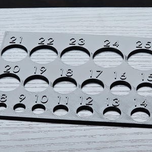 May include: A gray plastic ring sizing gauge with numbered circles from 8 to 27. The gauge is used to determine the size of rings.