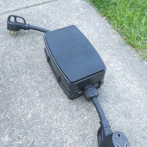 May include: Black outdoor electrical outlet extension box with a black cord and two standard three-pin plugs.
