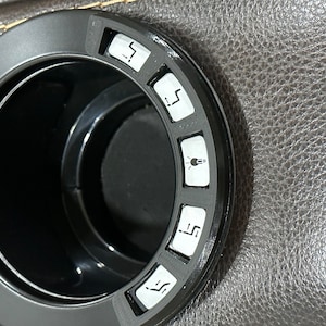 May include: A black cup holder with five buttons around the edge. The buttons have symbols for reclining, heat, and a light.