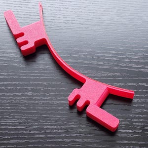 May include: A bright pink plastic phone stand with a curved design. The stand has two prongs on each end to hold a phone in place.