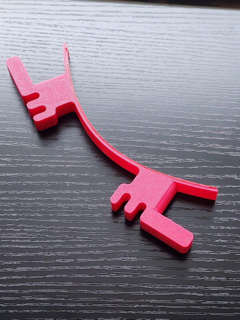 May include: A bright pink plastic phone stand with a curved design. The stand has two prongs on each end to hold a phone in place.