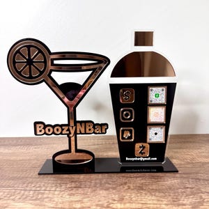 May include: A decorative bar sign featuring a cocktail glass and a cocktail shaker. The sign is made of copper-colored material with the text "BoozyNBar" and social media icons. The sign is on a black base and sits on a wooden surface.