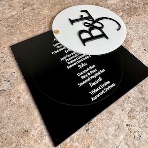 May include: A black and white menu with a circular design. The menu features a white circular top with the letters "B & F" in black. The menu lists dishes such as Jerk Chicken, Coconut Rice, and Assorted Sorbets.