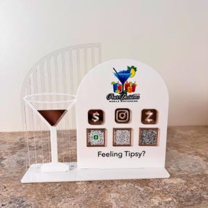 May include: A white acrylic mobile bartending display with a martini glass and a curved, slotted accent. The display features the text "Pour Decisions Mobile Bartending" and social media icons with QR codes. The phrase "Feeling Tipsy?" is also displayed.