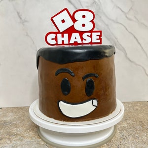 Peut inclure: Un g&acirc;teau au chocolat d&eacute;cor&eacute; d'un visage brun et d'un sourire blanc. Le g&acirc;teau est surmont&eacute; d'un topper rouge et blanc sur le th&egrave;me de Roblox qui dit "8 CHASE".