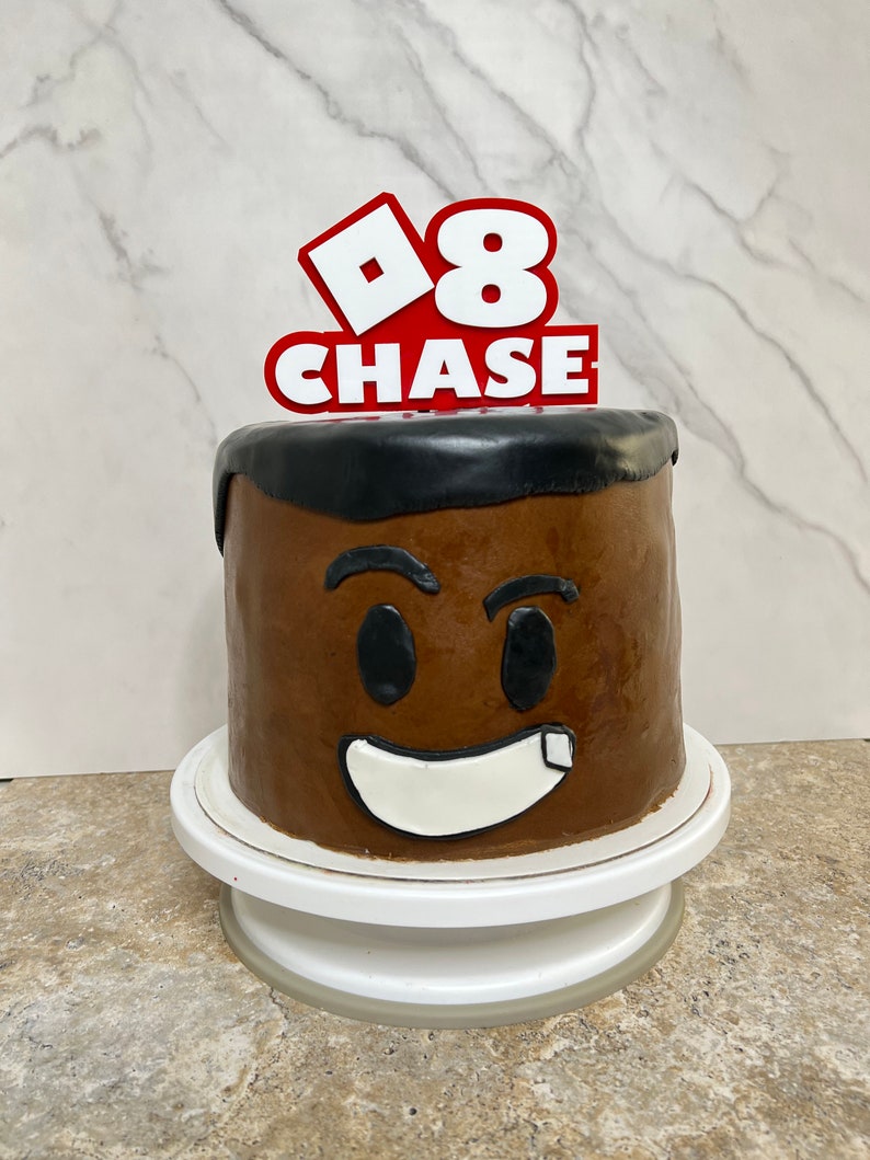 Peut inclure: Un g&acirc;teau au chocolat d&eacute;cor&eacute; d'un visage brun et d'un sourire blanc. Le g&acirc;teau est surmont&eacute; d'un topper rouge et blanc sur le th&egrave;me de Roblox qui dit "8 CHASE".