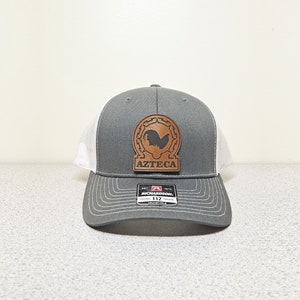 May include: A gray and white trucker hat with a brown leather patch featuring a rooster silhouette and the text "AZTECA".