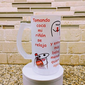 May include: White ceramic mug with a red and black Coca-Cola logo and a cartoon character holding a bottle of Coca-Cola. The mug has red text that reads "Tomando coca mi riñón se relaja y entre más bebo, menos quiero agua."