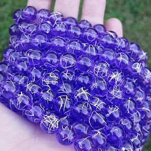 May include: A strand of translucent purple beads with white, web-like designs. The beads are round and held in a hand against a blurred green background. The beads are likely for jewelry making.