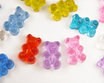 May include: A collection of translucent gummy bear-shaped charms in various colors, including red, purple, pink, blue, and yellow. The charms are scattered on a white surface, creating a playful and colorful arrangement. These plastic charms are ideal for crafting.