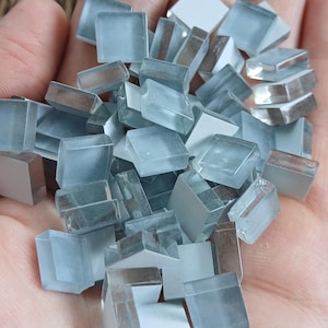 May include: A handful of small, square, light blue glass tiles. The tiles are a translucent, frosted color and appear to be for mosaic art or craft projects. Some tiles have a reflective, mirrored backing.
