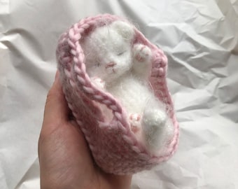 May include: A small white crocheted animal, resembling a baby bear, is sleeping inside a pink crocheted blanket.