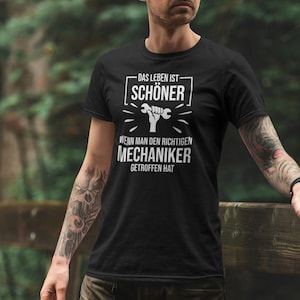 Puede incluir: Camiseta negra con texto blanco: "DAS LEBEN IST SCHÖNER" sobre un gráfico de llave inglesa. Debajo, el texto continúa: "WENN MAN DEN RICHTIGEN MECHANIKER GETROFFEN HAT". La camiseta es de cuello redondo.