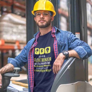 Puede incluir: Un hombre con un casco amarillo y una camisa vaquera azul se encuentra frente a una carretilla elevadora. Lleva una camiseta azul marino con el texto "Ich bin Elektriker Ich löse Probleme von denen du keine Ahnung hast auf eine Weise, die du nie verstehen wirst".