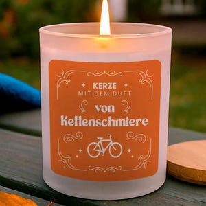 May include: A lit, frosted glass candle with an orange label. The label features the text "KERZE MIT DEM DUFT von Kettenschmiere" and a bicycle graphic. The candle sits on a wooden surface, with a wooden lid visible.