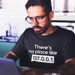 Può includere: Una persona indossa una maglietta nera con la scritta "There's no place like 127.0.0.1" stampata su di essa. La persona sta guardando un laptop.