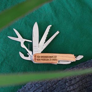 Puede incluir: Un cuchillo multiusos de bolsillo con mango de madera grabado con el texto "Für unseren Papa ist kein Problem zu groß". El cuchillo tiene una variedad de herramientas, incluyendo una hoja, una sierra, un destornillador y un abridor de botellas.
