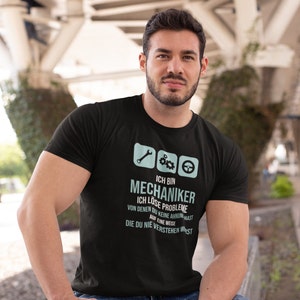 Puede incluir: Una camiseta negra con un gráfico azul de una llave inglesa, engranajes y un volante. El texto en la camiseta dice "Ich bin Mechaniker. Ich löse Probleme von denen du keine Ahnung hast auf eine Weise die du nie verstehen wirst."