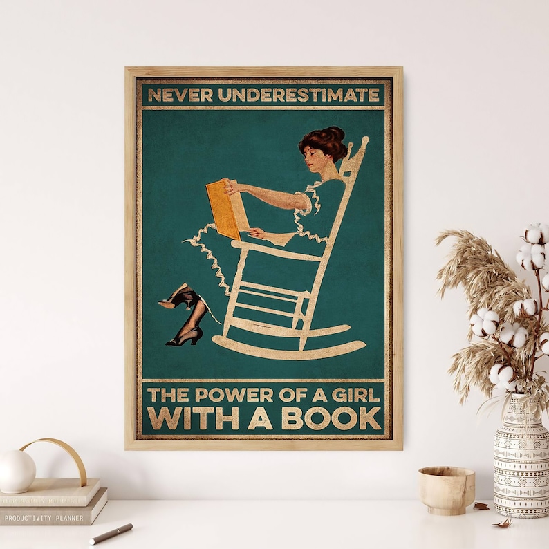 May include: A framed vintage-style poster with the text "NEVER UNDERESTIMATE THE POWER OF A GIRL WITH A BOOK." The poster features a woman in a rocking chair reading a book, with a teal background and gold accents. The frame is a light brown color.