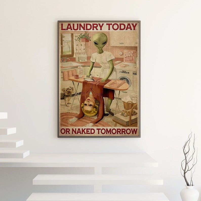 May include: A framed poster with the text "LAUNDRY TODAY OR NAKED TOMORROW." The image depicts an alien ironing a woman upside down. A dog and laundry room appliances are in the background. The poster has a vintage, retro style.