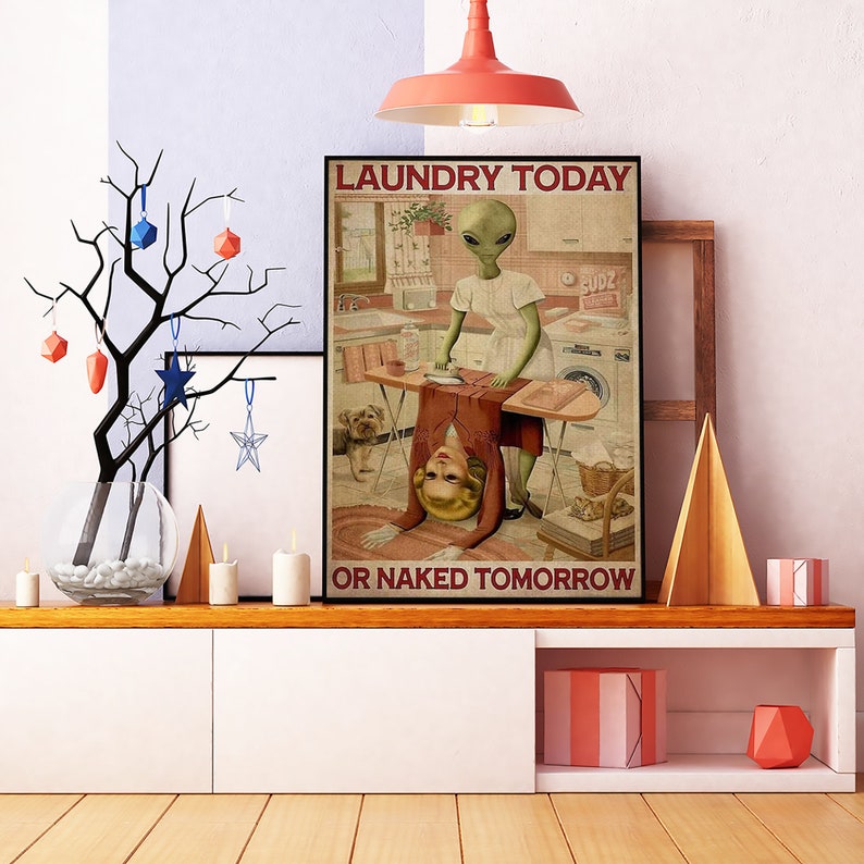 May include: A framed poster with the text "LAUNDRY TODAY OR NAKED TOMORROW" features an alien ironing a red dress while a woman is bent over. The background shows a kitchen with a washing machine and a dog.