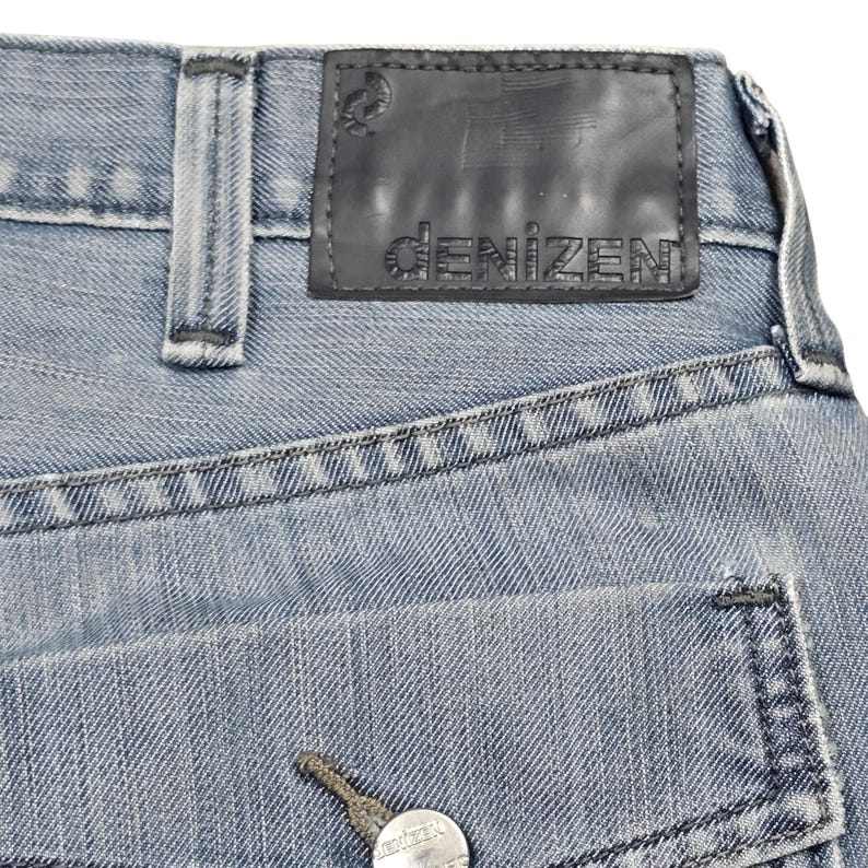 Puede incluir: Primer plano de unos vaqueros de mezclilla azul claro. Los vaqueros tienen un parche de cuero negro con el nombre de la marca "denizen" y un peque&ntilde;o logotipo. El bolsillo trasero es visible, junto con los detalles de los botones y las costuras.