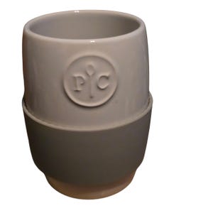 Puede incluir: Una taza de cerámica gris con una banda de silicona gris alrededor de la base. La taza tiene un logotipo circular en relieve con las letras "P" y "C". La taza tiene una forma redondeada y una abertura ancha.