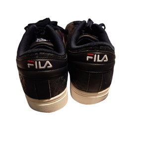 May include: A pair of black sneakers with white soles. The heels feature the brand name "FILA" in white lettering with a red accent. The shoes have a textured pattern and dark blue laces.