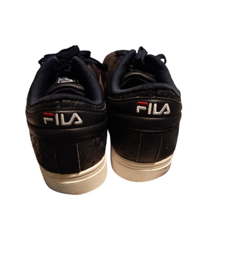 May include: A pair of black sneakers with white soles. The heels feature the brand name "FILA" in white lettering with a red accent. The shoes have a textured pattern and dark blue laces.