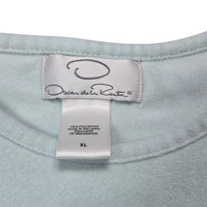 Op de afbeelding: Close-up van een lichtblauw kledingstuk met een label met het "Oscar de la Renta" logo. Het label geeft aan dat het materiaal 100% polyester is, gemaakt in Sri Lanka en maat XL.