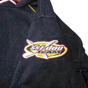 May include: Black fleece jacket with a "Ski-Doo TEAM" patch on the sleeve. The patch features a yellow and white design with the brand name in red and white. The jacket has a high collar and a yellow accent.