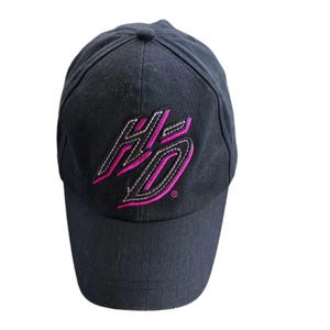 May include: A black baseball cap featuring an embroidered logo on the front panel. The logo consists of the letters HD in a stylized font, outlined in white stitching with a vibrant pink fill. The cap has a structured crown and a curved brim.