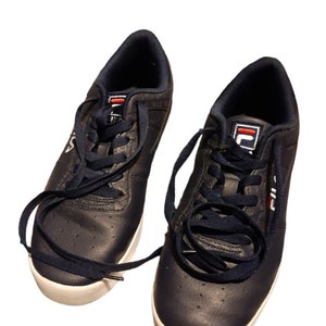 May include: A pair of navy blue Fila sneakers with white soles and navy blue laces. The sneakers have the Fila logo in white and red on the tongue and side. The shoes are made of a leather-like material.
