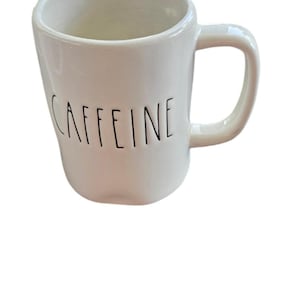Puede incluir: Taza de cerámica blanca con cuerpo curvo y asa. La palabra "CAFFEINE" está impresa en negro, en mayúsculas, en la parte delantera de la taza. La taza está diseñada para bebidas calientes.