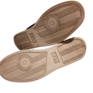 May include: Two athletic shoes with tan soles and black uppers are displayed. The soles feature a textured pattern with the brand name "FILA" embossed on the side. The shoes are set against a white background.