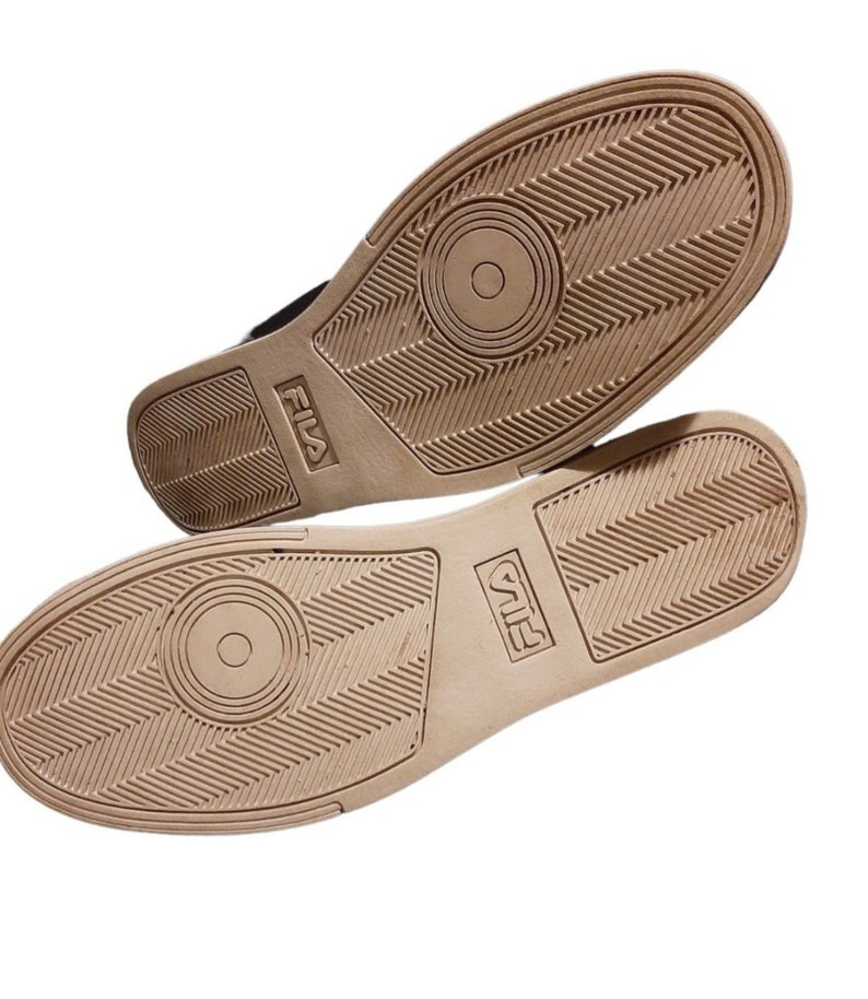 May include: Two athletic shoes with tan soles and black uppers are displayed. The soles feature a textured pattern with the brand name "FILA" embossed on the side. The shoes are set against a white background.