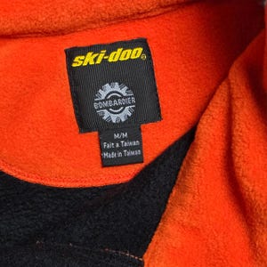 May include: Close-up of an orange and black fleece jacket. The jacket features a black tag with the "ski-doo" logo and "BOMBARDIER" text. The tag also includes "M/M", "Fait a Taiwan", and "Made in Taiwan" text.