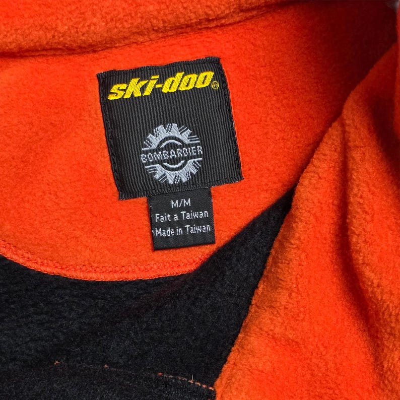 May include: Close-up of an orange and black fleece jacket. The jacket features a black tag with the "ski-doo" logo and "BOMBARDIER" text. The tag also includes "M/M", "Fait a Taiwan", and "Made in Taiwan" text.